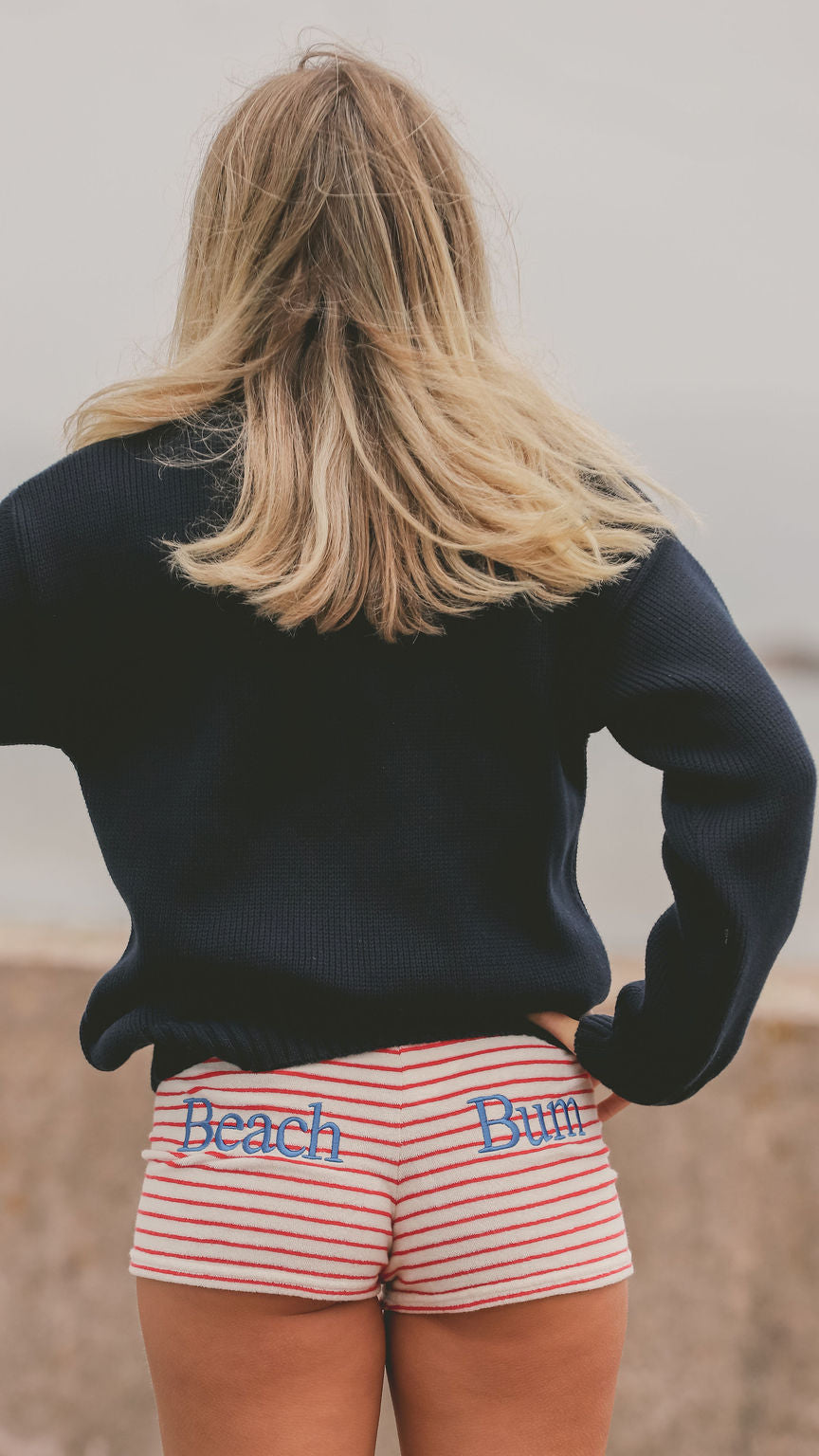 A woman shown from behind wearing a dark navy long-sleeve top and red-and-white striped shorts with the words 'Beach Bum' embroidered in blue on the back.