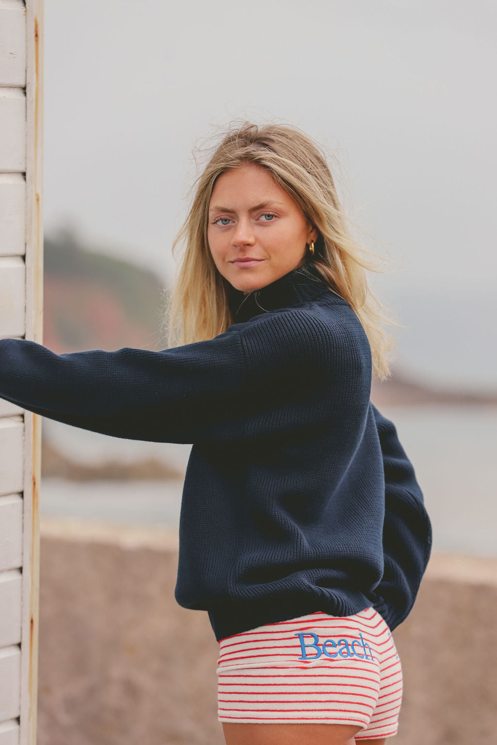 A woman wearing a navy blue ribbed turtleneck sweater and red and white striped shorts with the word 'Beach' embroidered in blue, leaning against a white brick wall outdoors.